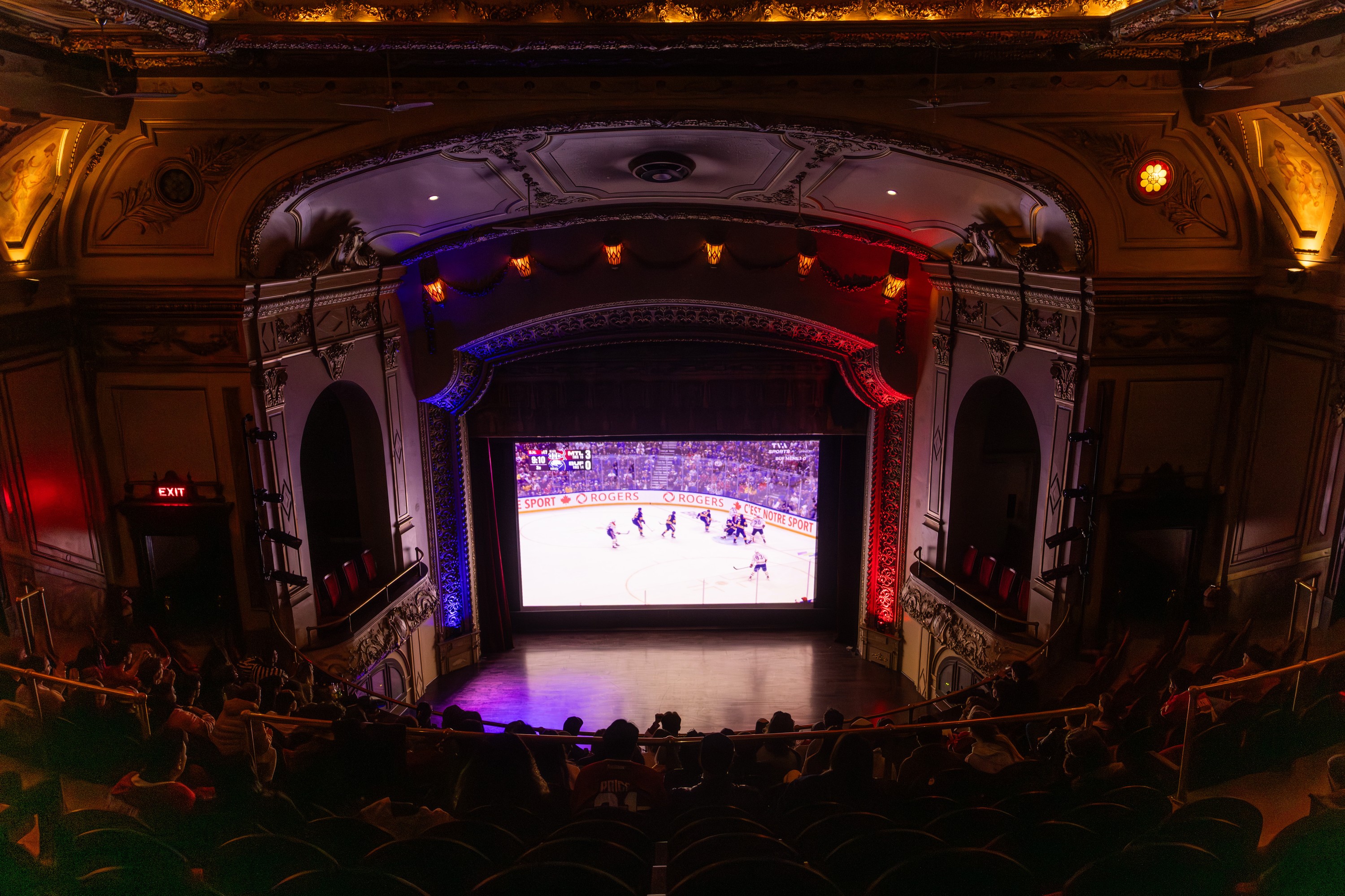 A hockey game is projected on the screen of an old-time theatre with a packed audience