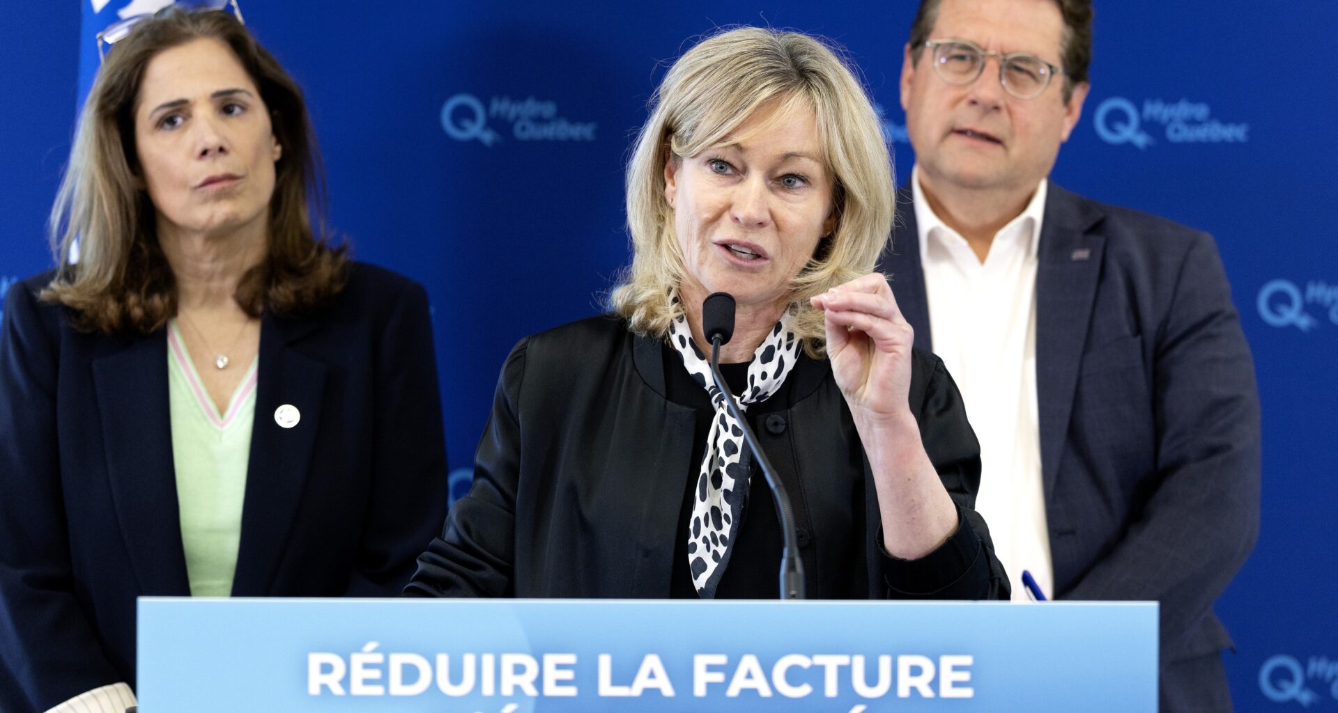 Claudine Bouchard speaks at a podium that has 'Réduire la facture d'électricité' on it, in front of Pascal Déry and Bernard Drainville and a backdrop with Hydro-Québec logos.