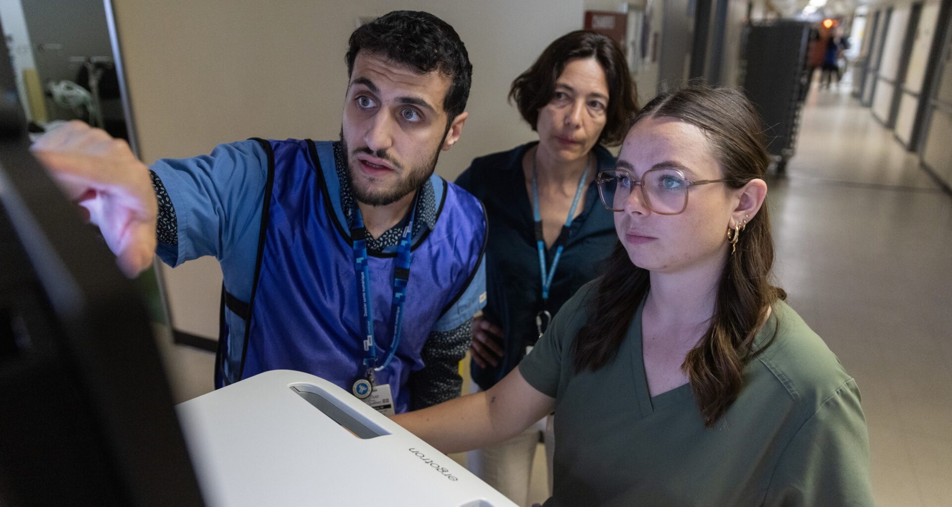 A man and two women stand in hospital corridor, looking at computer screen.