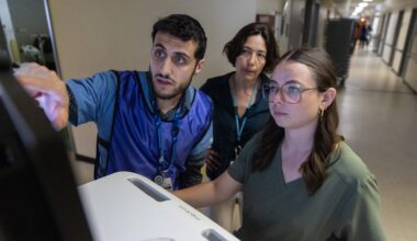 A man and two women stand in hospital corridor, looking at computer screen.