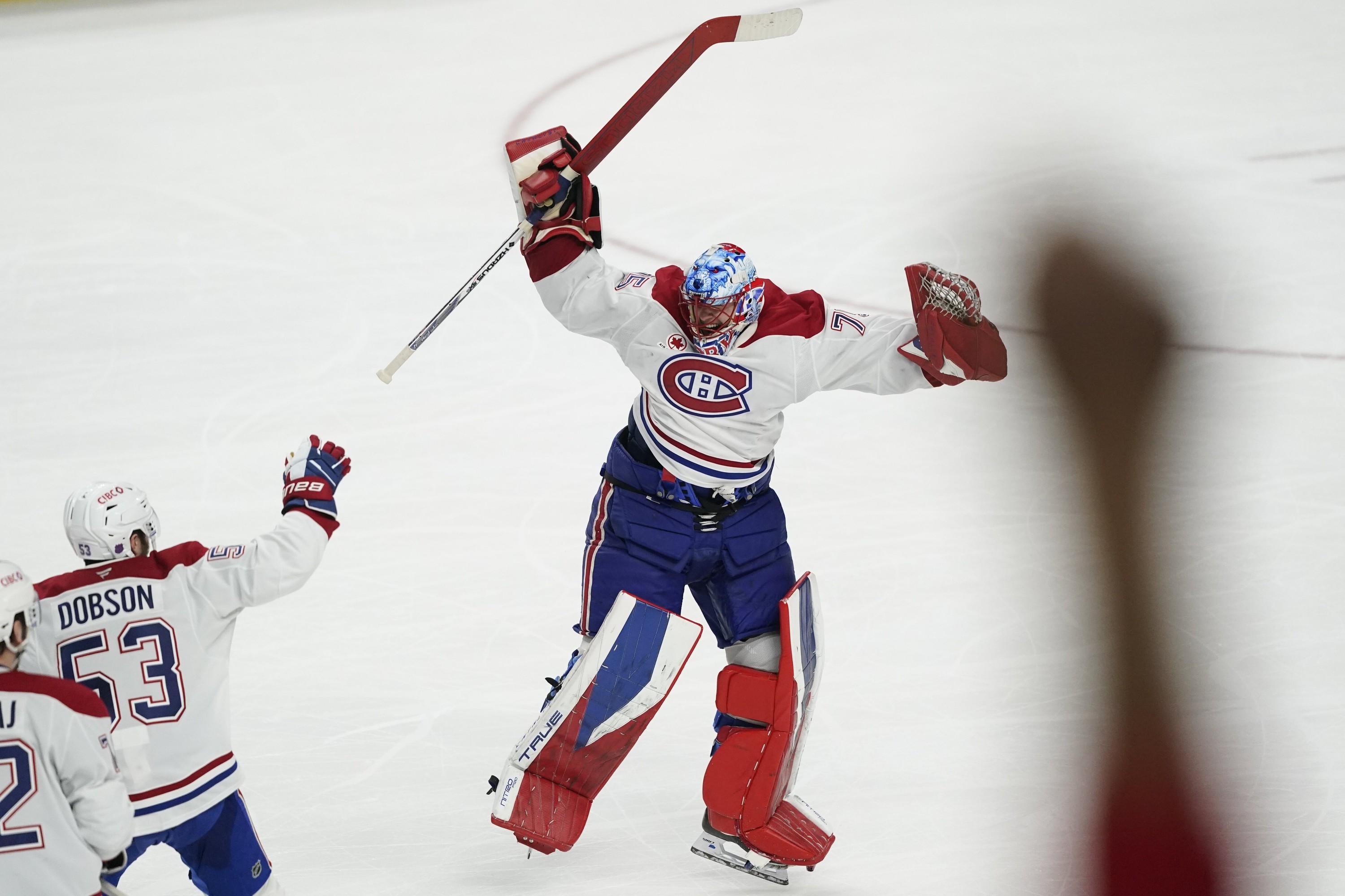 Montreal Canadiens goaltender Jakub Dobes jumps for joy on the ice as teammates skate toward him. In the foreground, a blurry fist rises into the air.