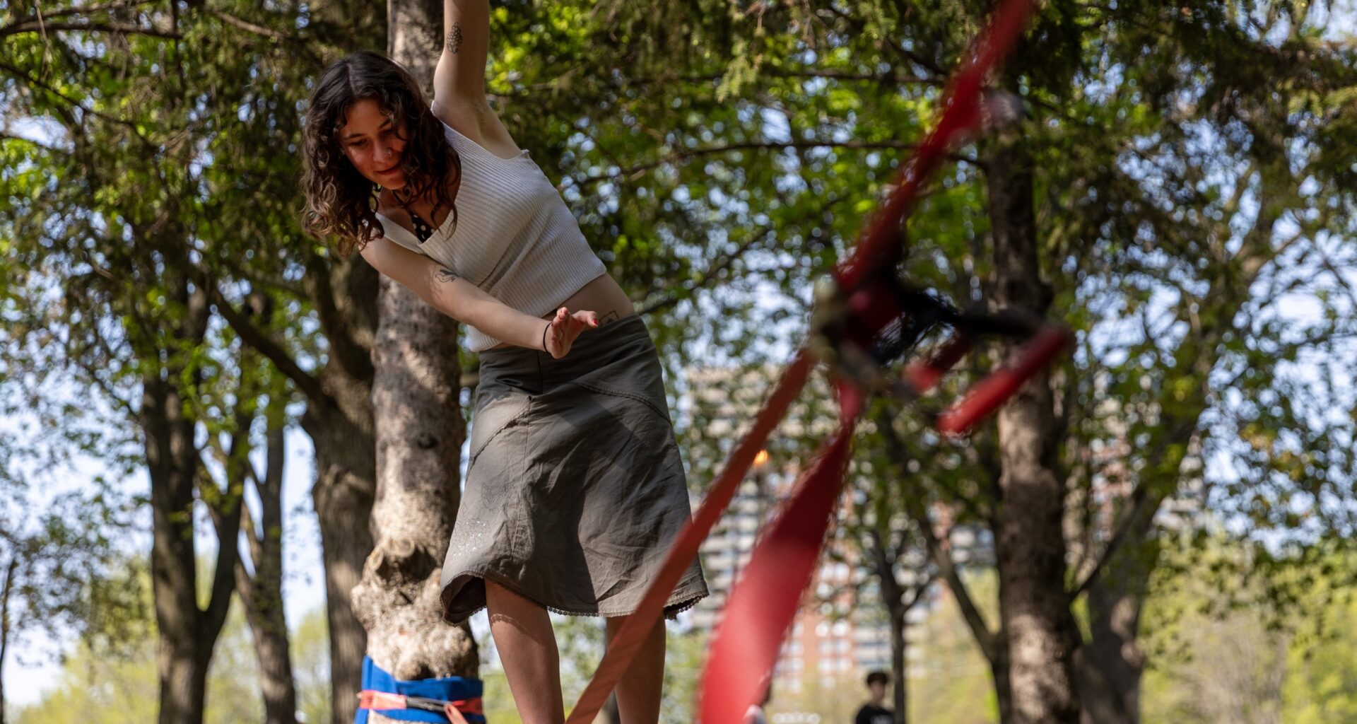 A woman balances on a tightrope held between two trees in a park.