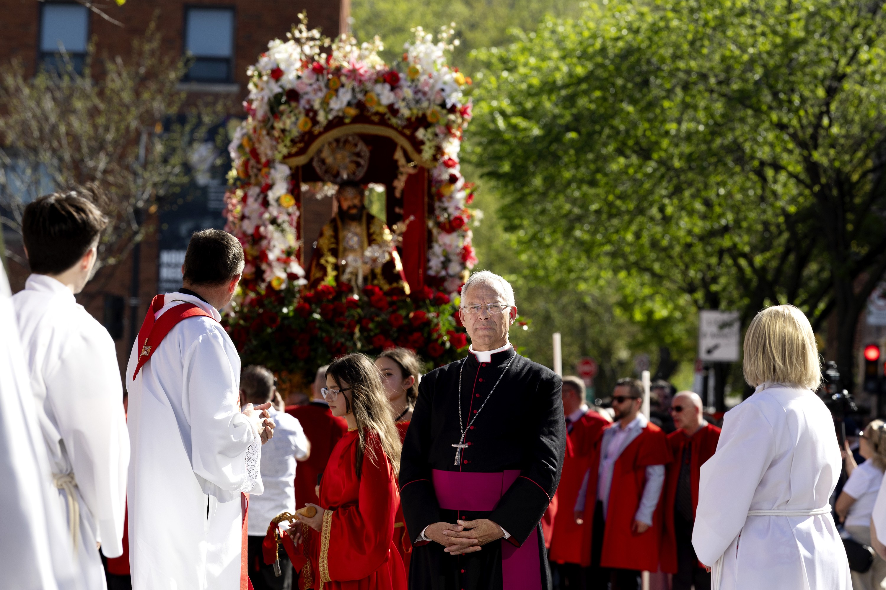 People in Catholic attire stand in front of a raised statue of Jesus decorated with flowers during an outdoor procession.