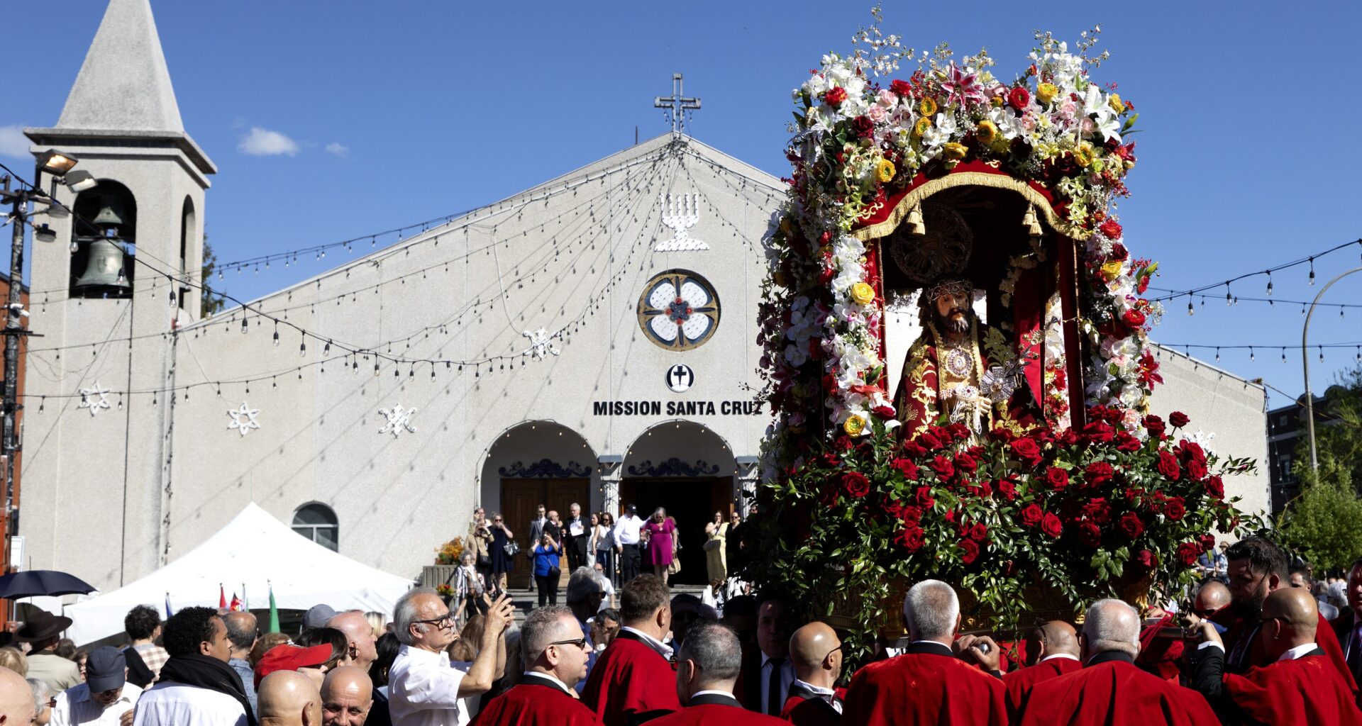 A crowd of people, some in religious garb, surround a statue of Jesus amid flowers that is being held aloft outside a church.
