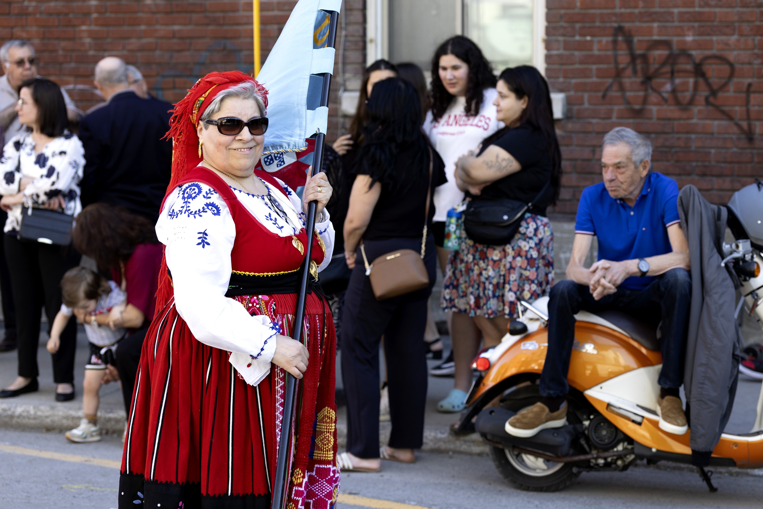 A woman smiles as she holds an obscured flag, with people milling about on the sidewalk behind her.