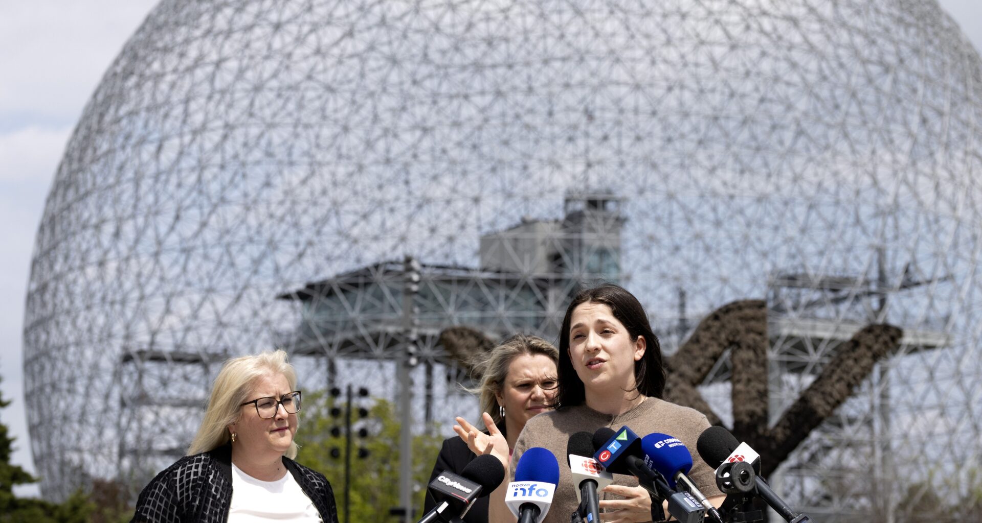 A woman speaks in front of a bank of media microphones as two other women listen behind her, as the trio stand in front of Montreal's Biosphère.