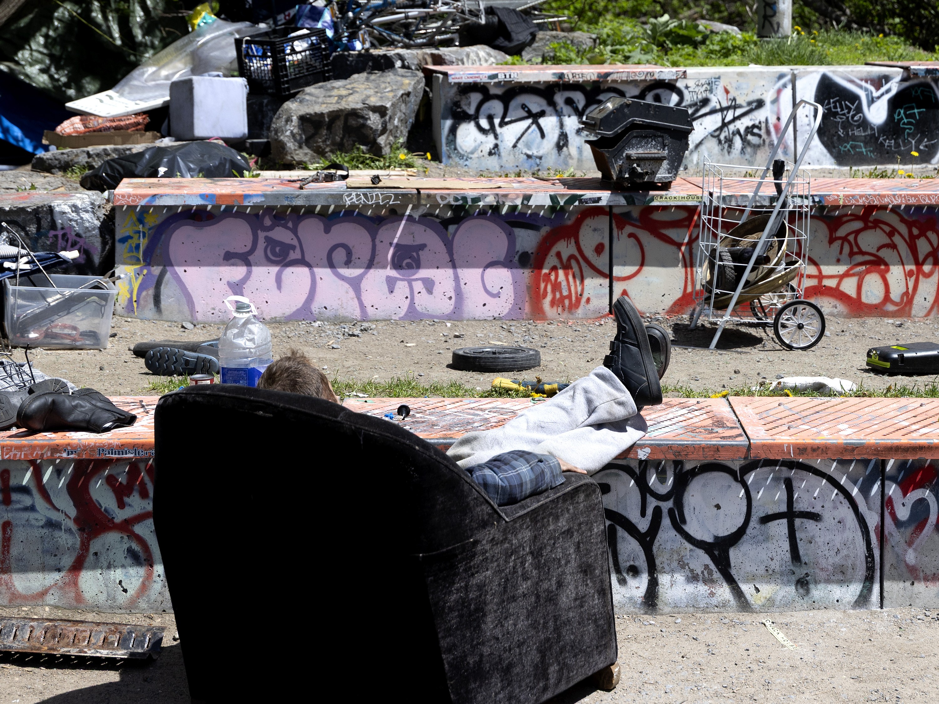 Seen from behind, a person sits in an armchair with their feet on a short concrete wall covered in grafitti. There are items around the area from a homeless encampment.