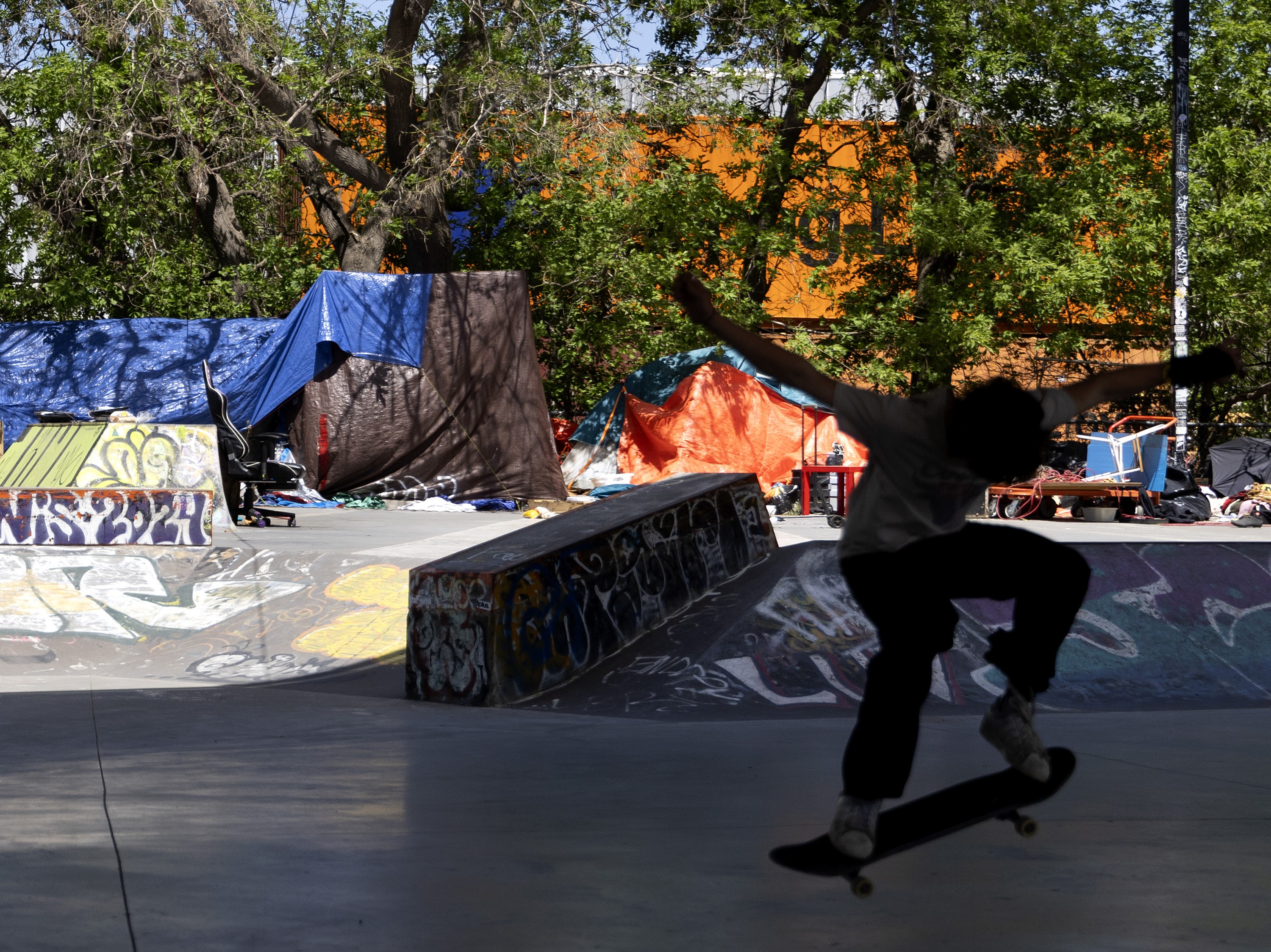 A person in silhouette skateboards in front of a homeless encampment.