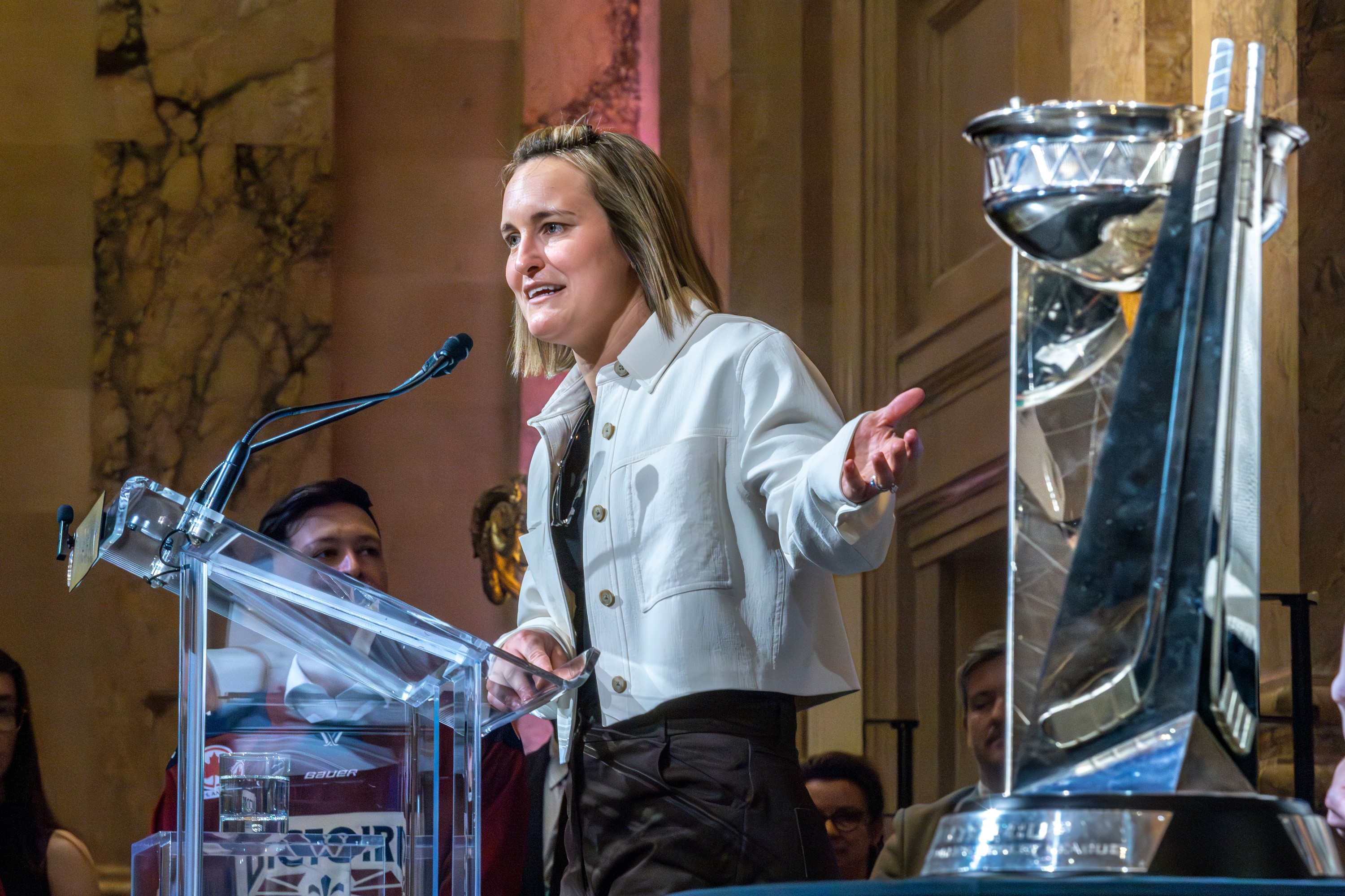 Woman stands at microphone, left arm pointing toward the Walter Cup on a table.
