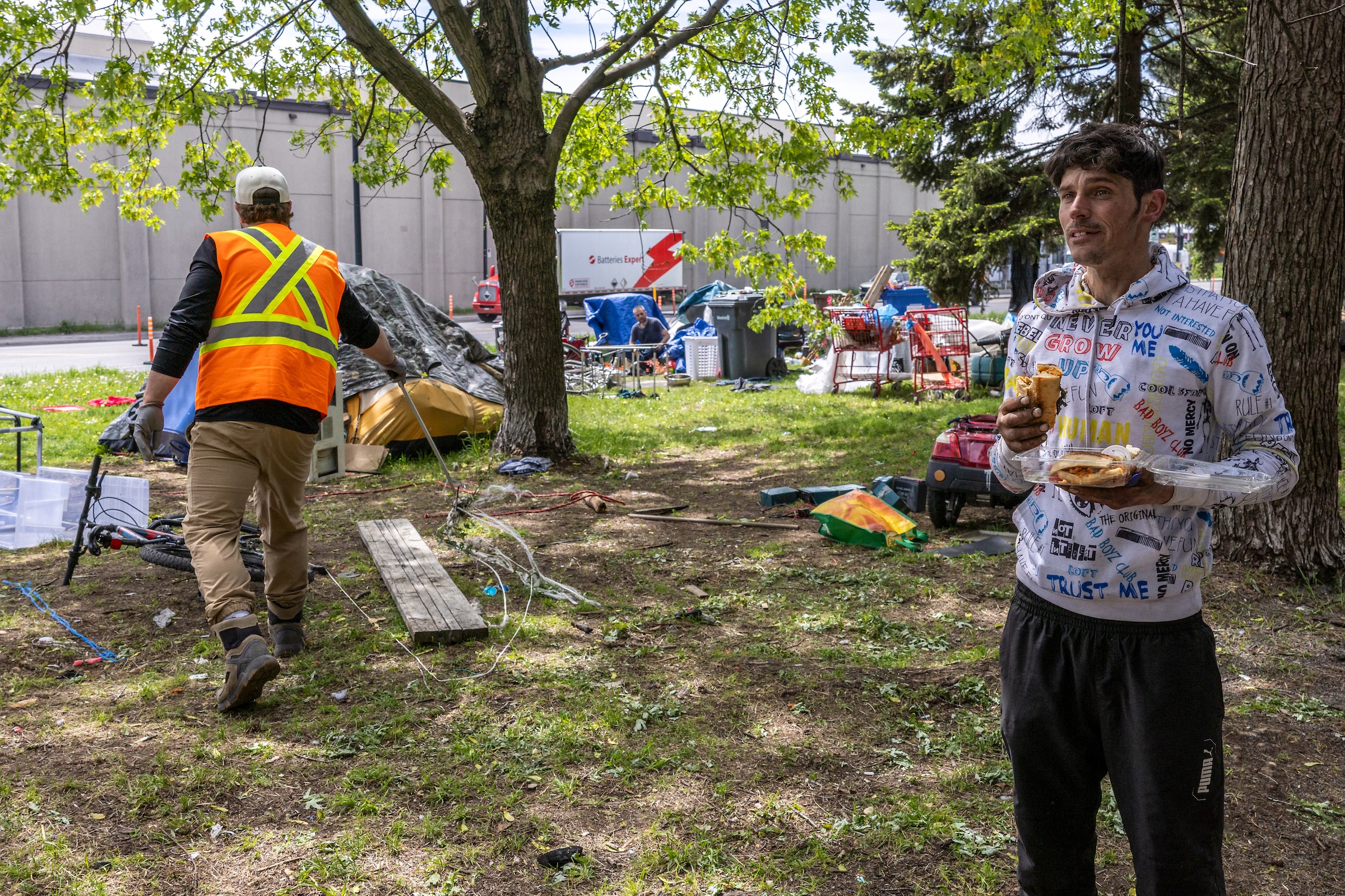A man eats standing on a green space while another man in a reflective vest does some work, with tents and various objects in the background.