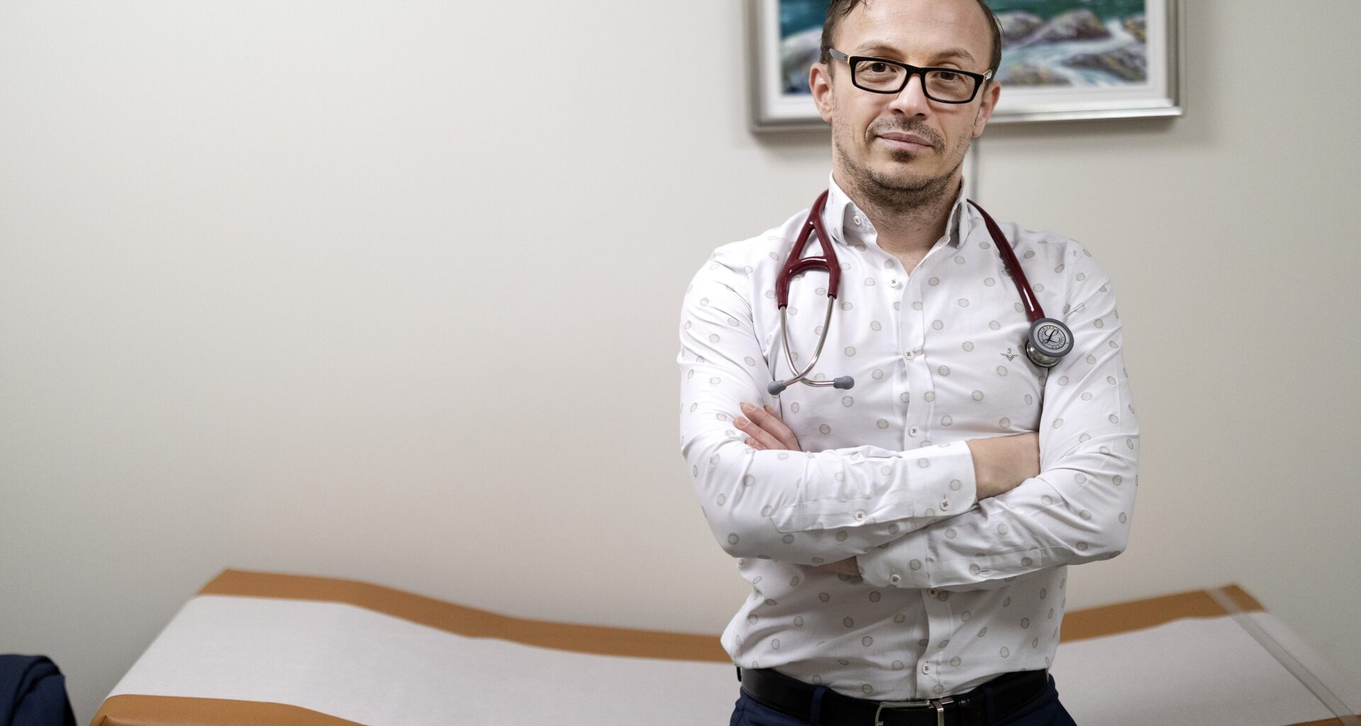 A doctor with a stethoscope around his neck crosses his arms as he leans against an examination table.