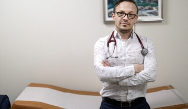 A doctor with a stethoscope around his neck crosses his arms as he leans against an examination table.