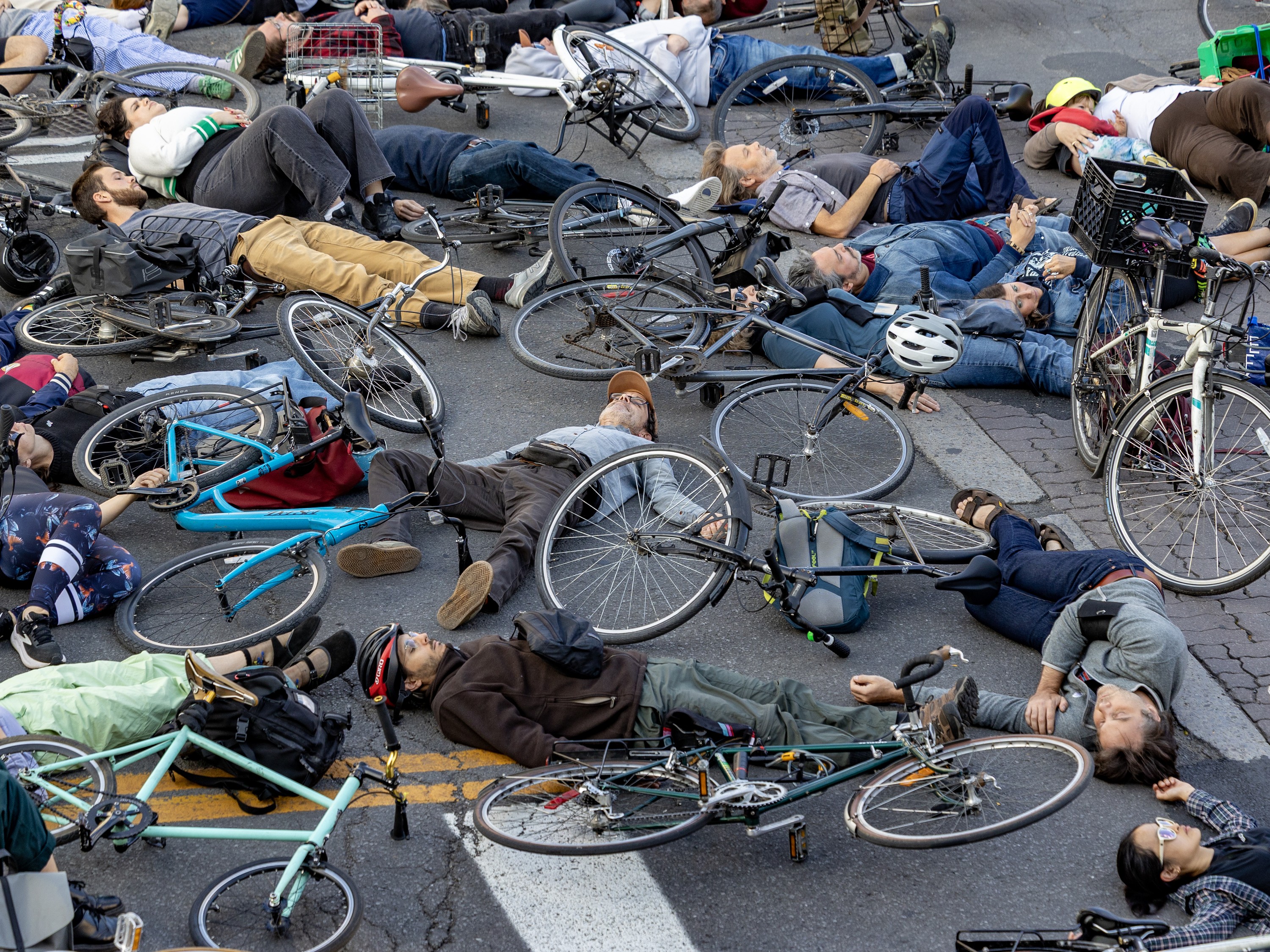 A group of cyclists lie next to their bikes on a city intersection, as though they have all been killed.