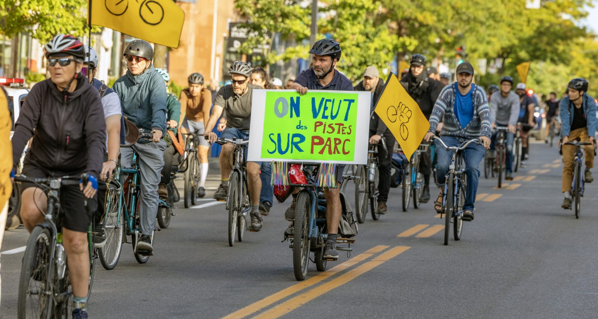 Cyclists on a city street, with one holding a placard that says On Veut des Pistes sur Parc.