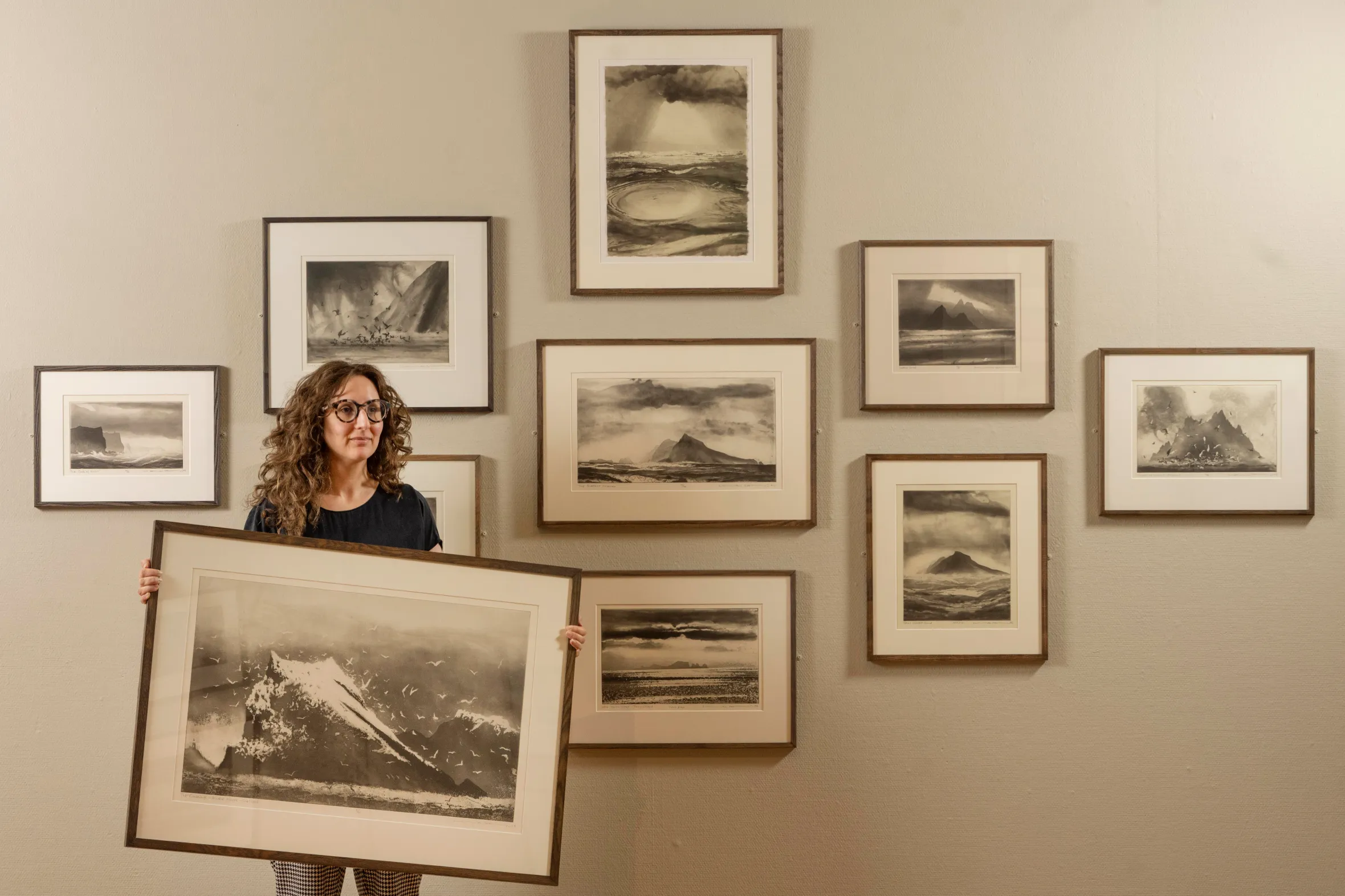 Curator Nathalie Levi holds a framed print of "The Rumblings" surrounded by other framed etchings by Norman Ackroyd, depicting British Isles' cliffs, shorelines and remote coasts.