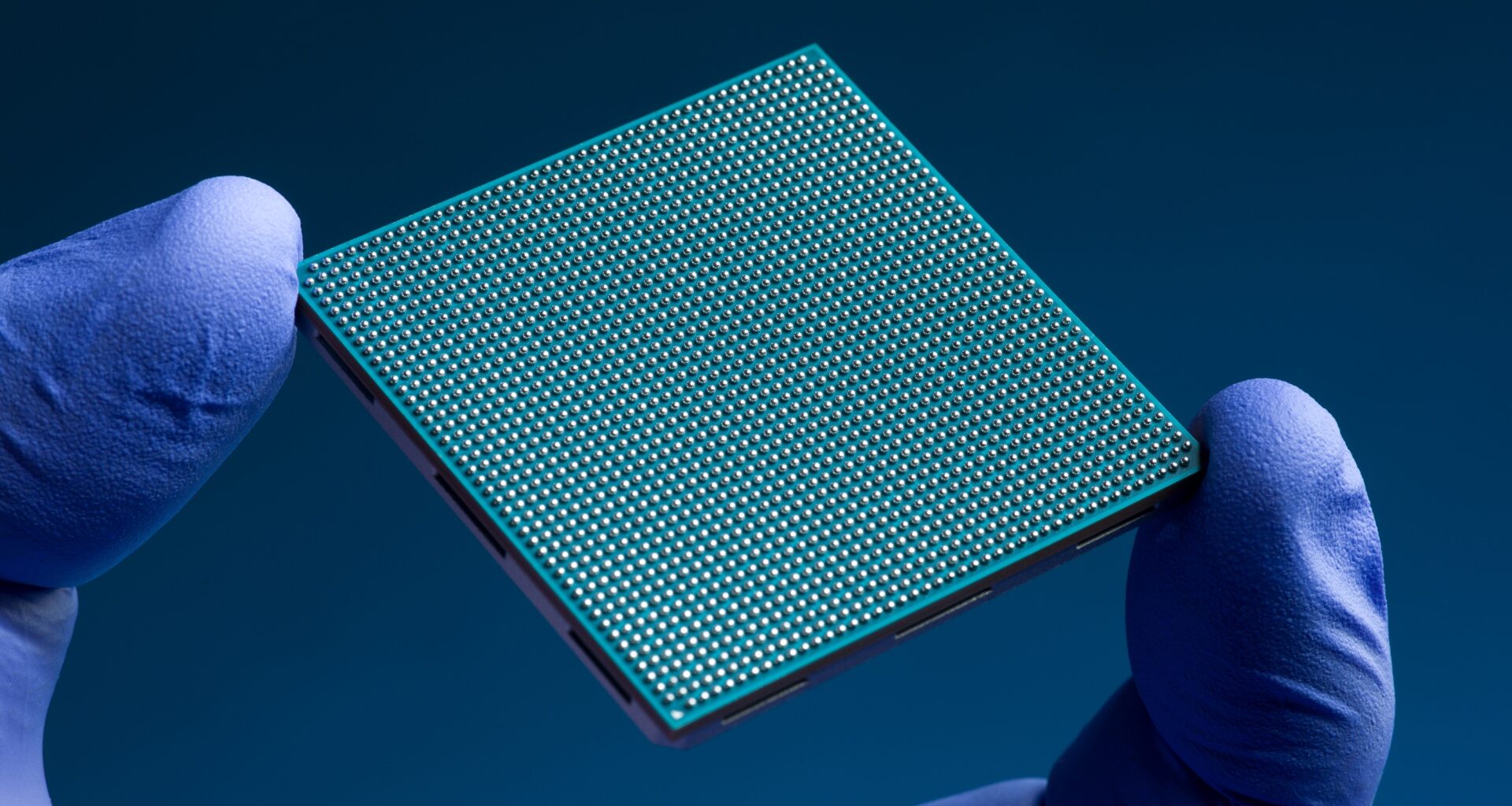 The finger and thumb of a hand clad in a blue nitrile glove hold a teal-colored square semiconductor chip against a dark blue background. The chip has an intricate grid of hundreds of tiny silver solder bumps on its underside.