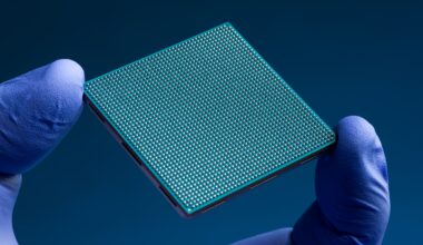 The finger and thumb of a hand clad in a blue nitrile glove hold a teal-colored square semiconductor chip against a dark blue background. The chip has an intricate grid of hundreds of tiny silver solder bumps on its underside.