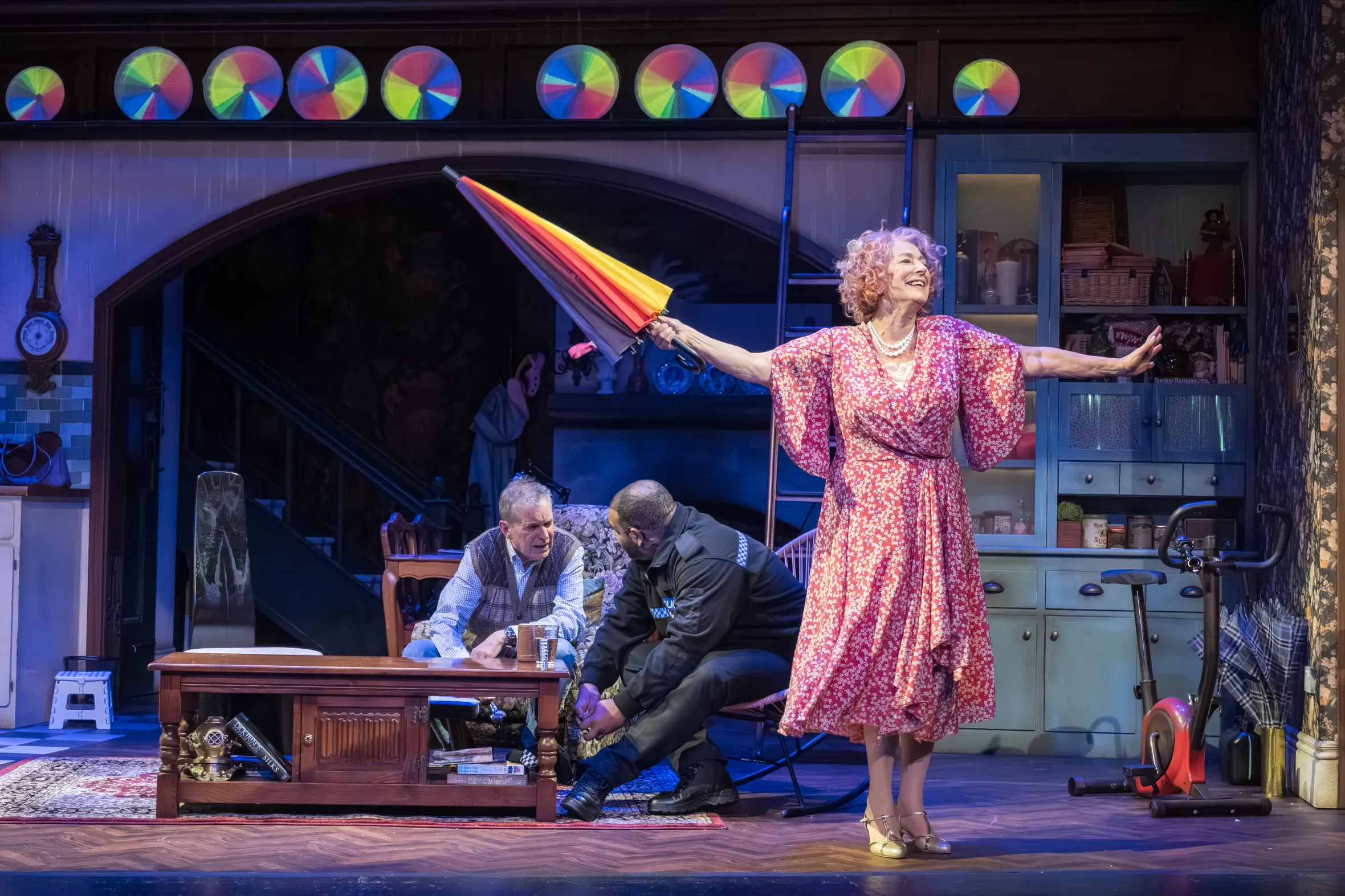 Maureen Lipman as Allegra holds an umbrella in a floral dress on stage.