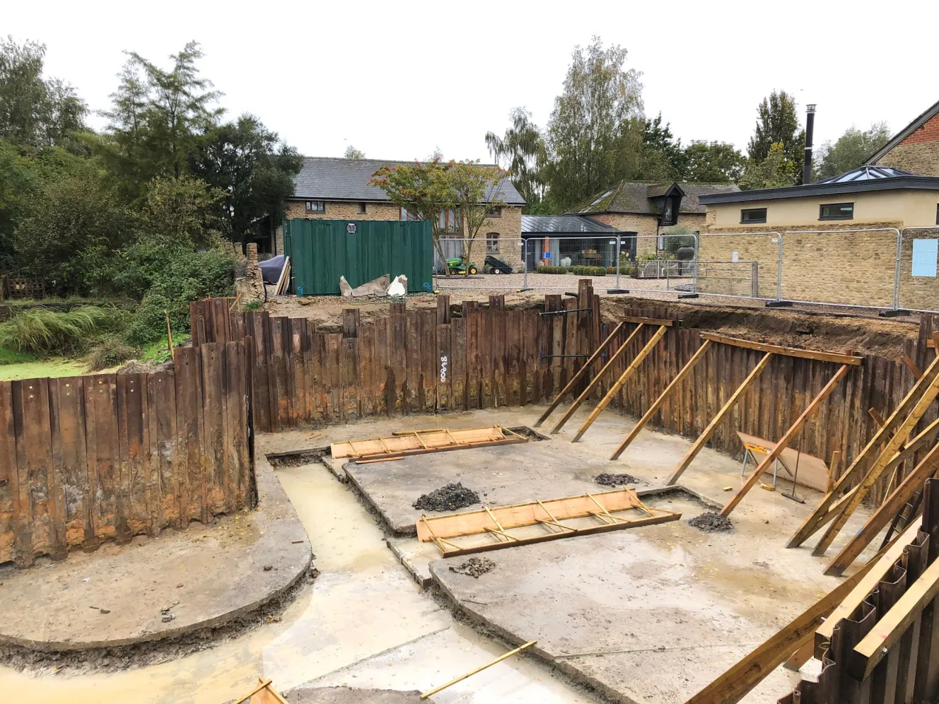 Construction site for a semi-subterranean home, with a deep excavation filled with rainwater, surrounded by sheet piling and wooden supports.
