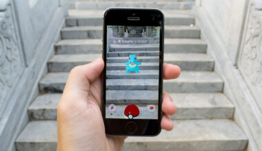 A hand holds a smartphone displaying the Pokémon Go app in augmented reality mode, showing the water-type Pokémon Totodile appearing on a set of stone steps.