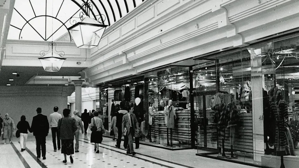 A black and white photo of the shopping centre with people dressed smartly walking down a corridor lined with shops. The central shop has manniquins in the window. The ceiling has an arched window.