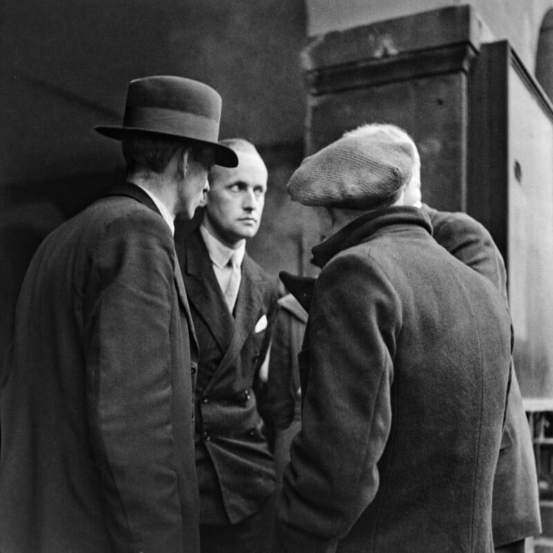 Four men stand in close conversation, dressed in coats and hats typical of early 20th-century fashion. Three have their backs to the camera, while one man faces forward, looking serious. The background is dark and indistinct.
