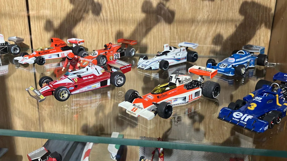 A glass display shelf holds detailed miniature Formula One race cars arranged neatly in rows.
The models feature bright liveries in red, white, blue, and orange, with visible sponsor logos and numbers.
Reflections on the glass and shadows on the wooden backing add depth to the carefully curated collection.