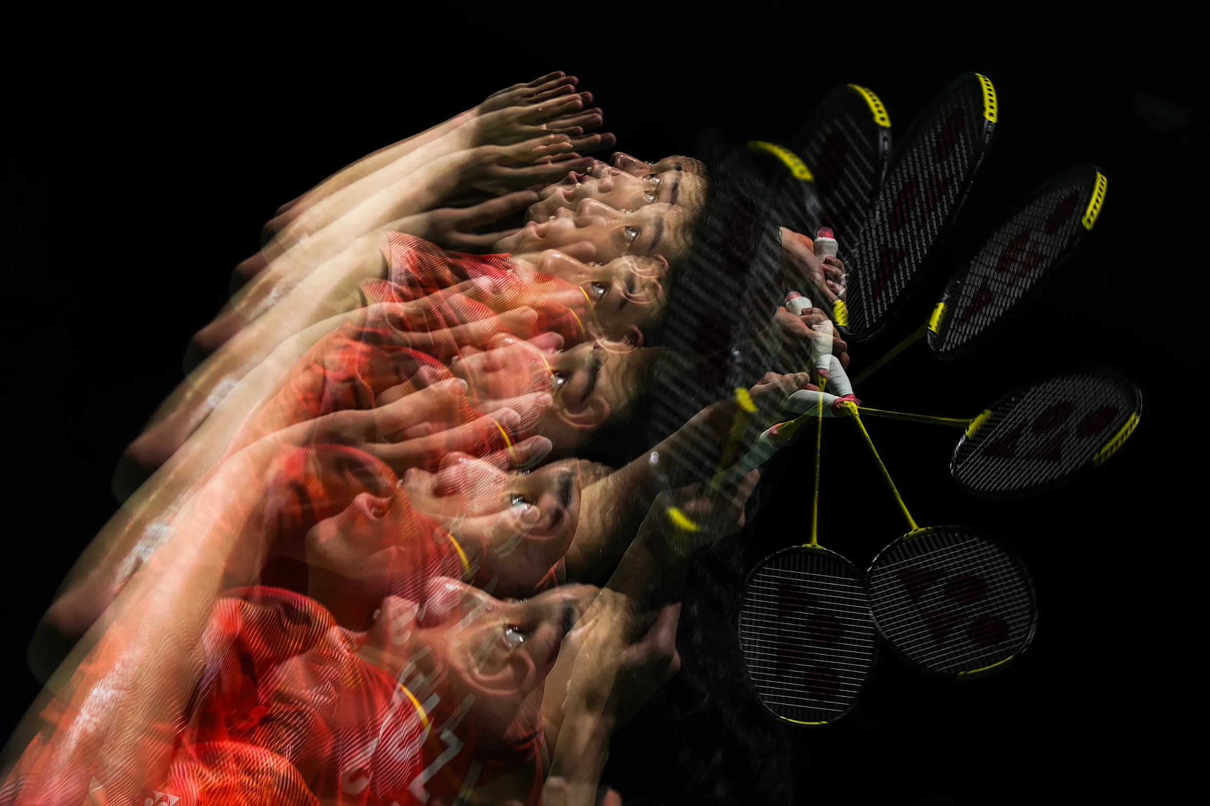 Multiple exposure image of a badminton player in a red shirt, shown as multiple transparent instances of himself, hitting a shuttlecock with a badminton racket.