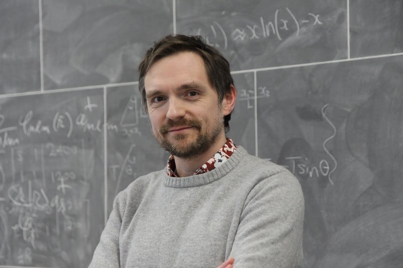 A medium shot of a smiling man with a beard and grey sweater standing in front of a chalkboard covered in mathematical equations.