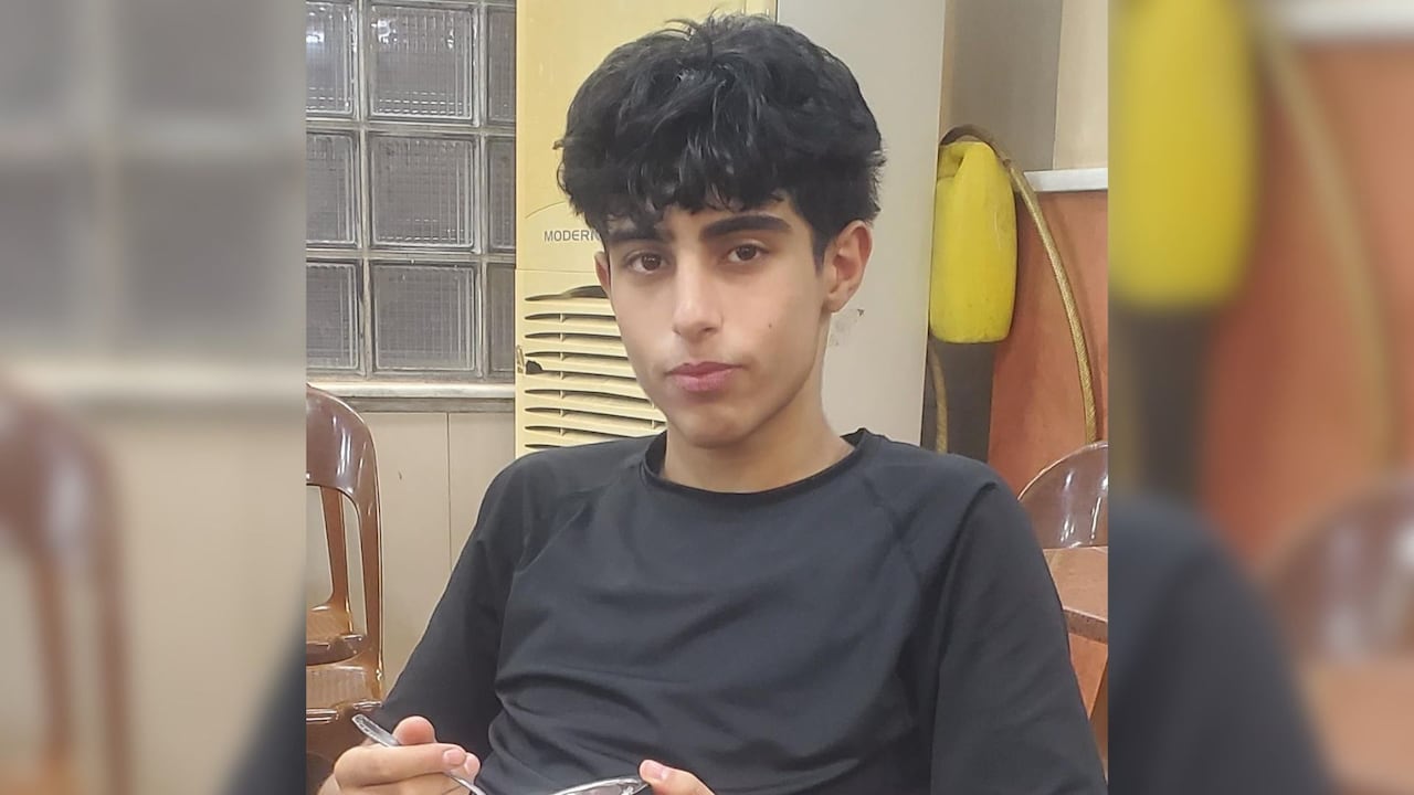 A portrait of a teenager with curly hair wearing a black T-shirt. He is seated at a table and holding a utensil and a plate.