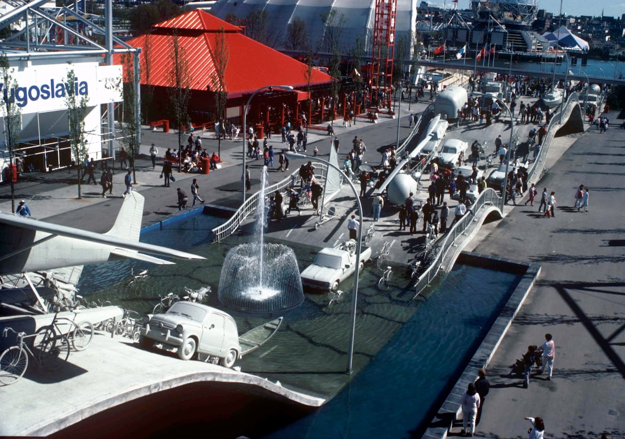People walk amidst a fantastical concrete roadway that crests and falls. The highway is covered in grey monochrome vehicles, including cars, trucks, planes and bicycles. A fountain is visible within the artwork.