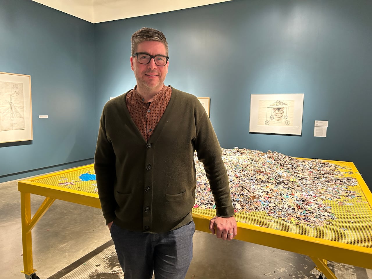 A man with glasses stands in front of a table covered in puzzle pieces.