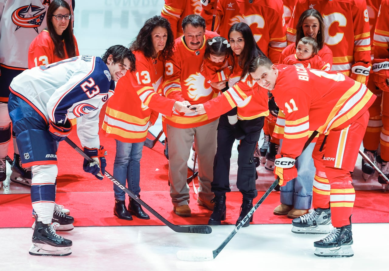 People of all ages in red hockey jerseys hold a puck over centre ice and two male players face off.