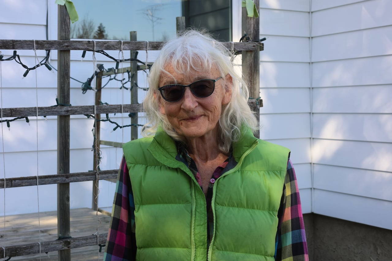 An elderly woman stands in front of a white house.