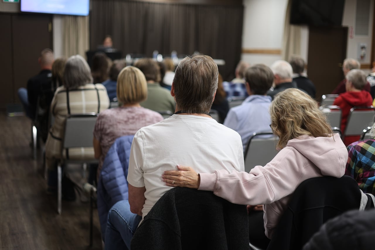A woman has her arm wrapped around a man as they sit amongst many people in a large meeting room.