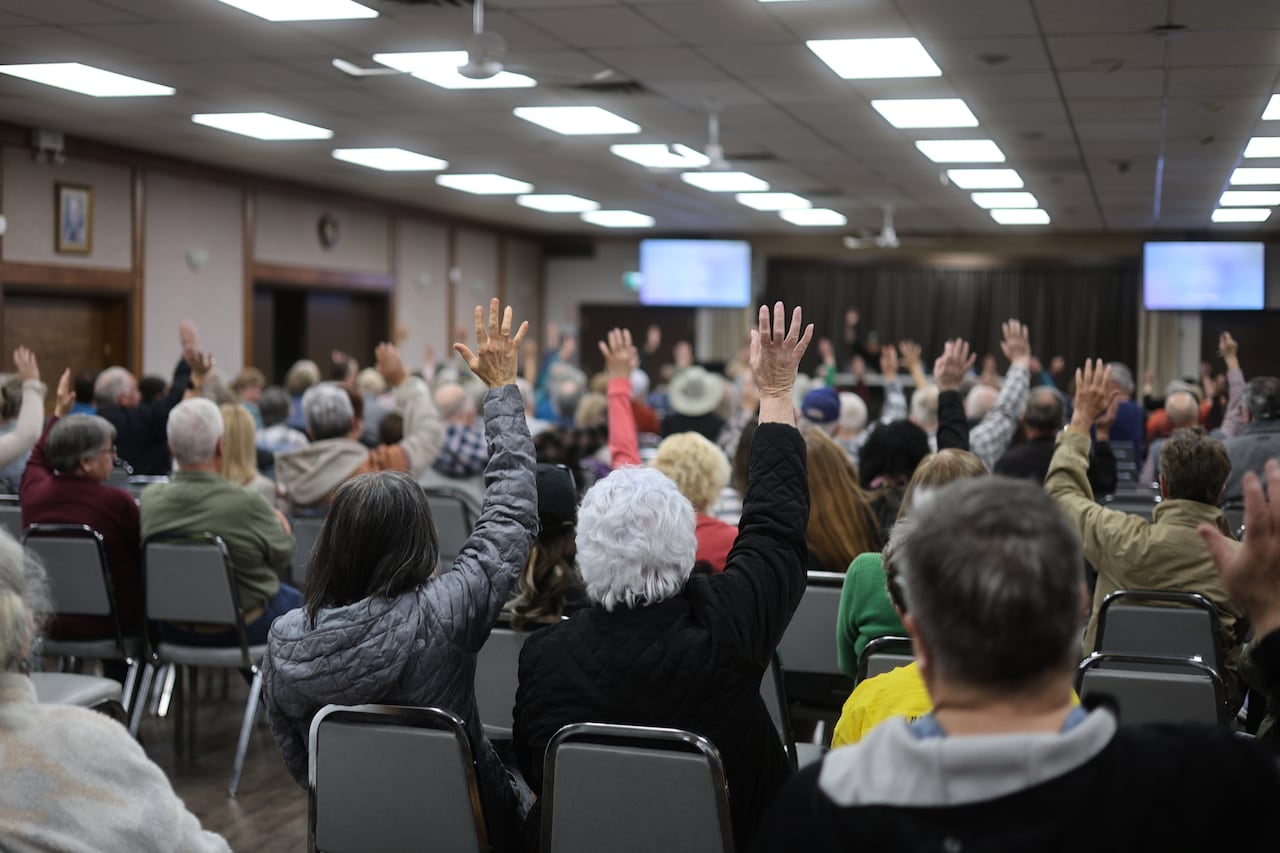 Several people seated in chairs in a meeting room raise their hands.