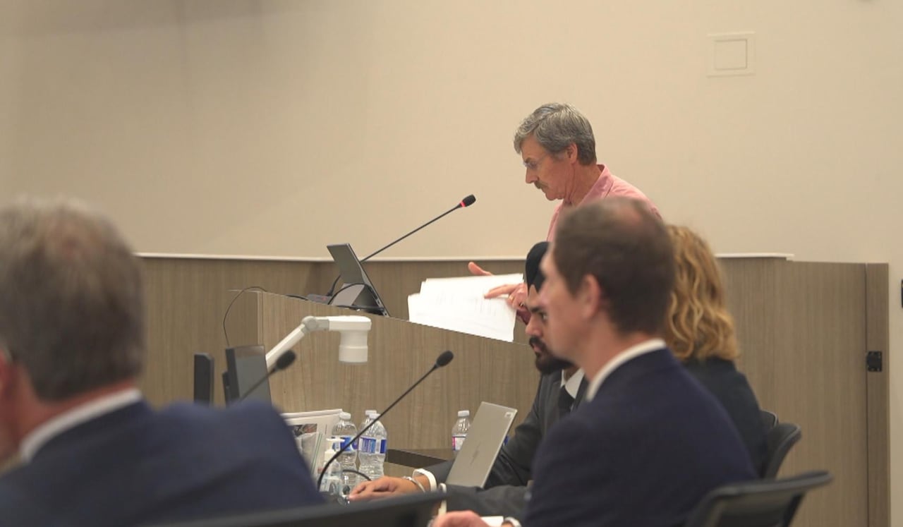A man rifles through papers as he stands at a podium in a meeting room.