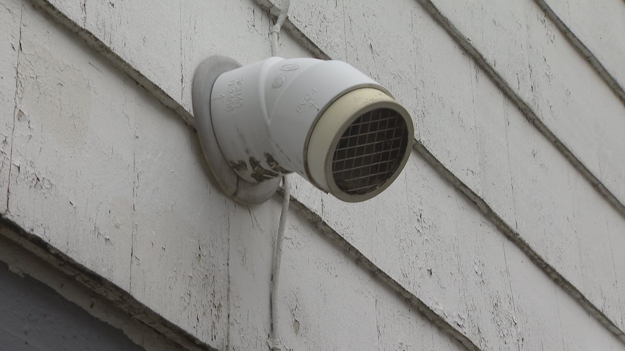 A pipe with a grate over the opening is seen coming out of an external wall of a building.