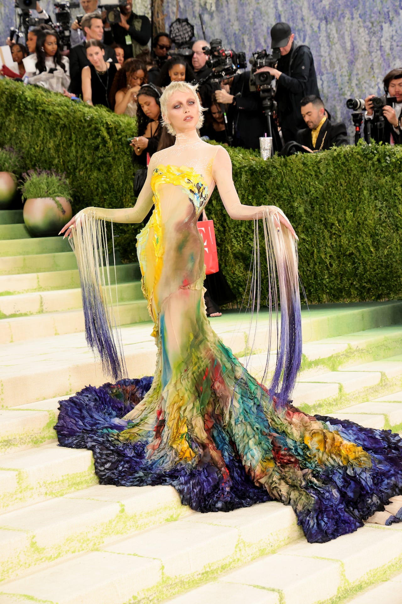A woman poses for a photo wearing a colourful gown with long fringe on both sleeves.