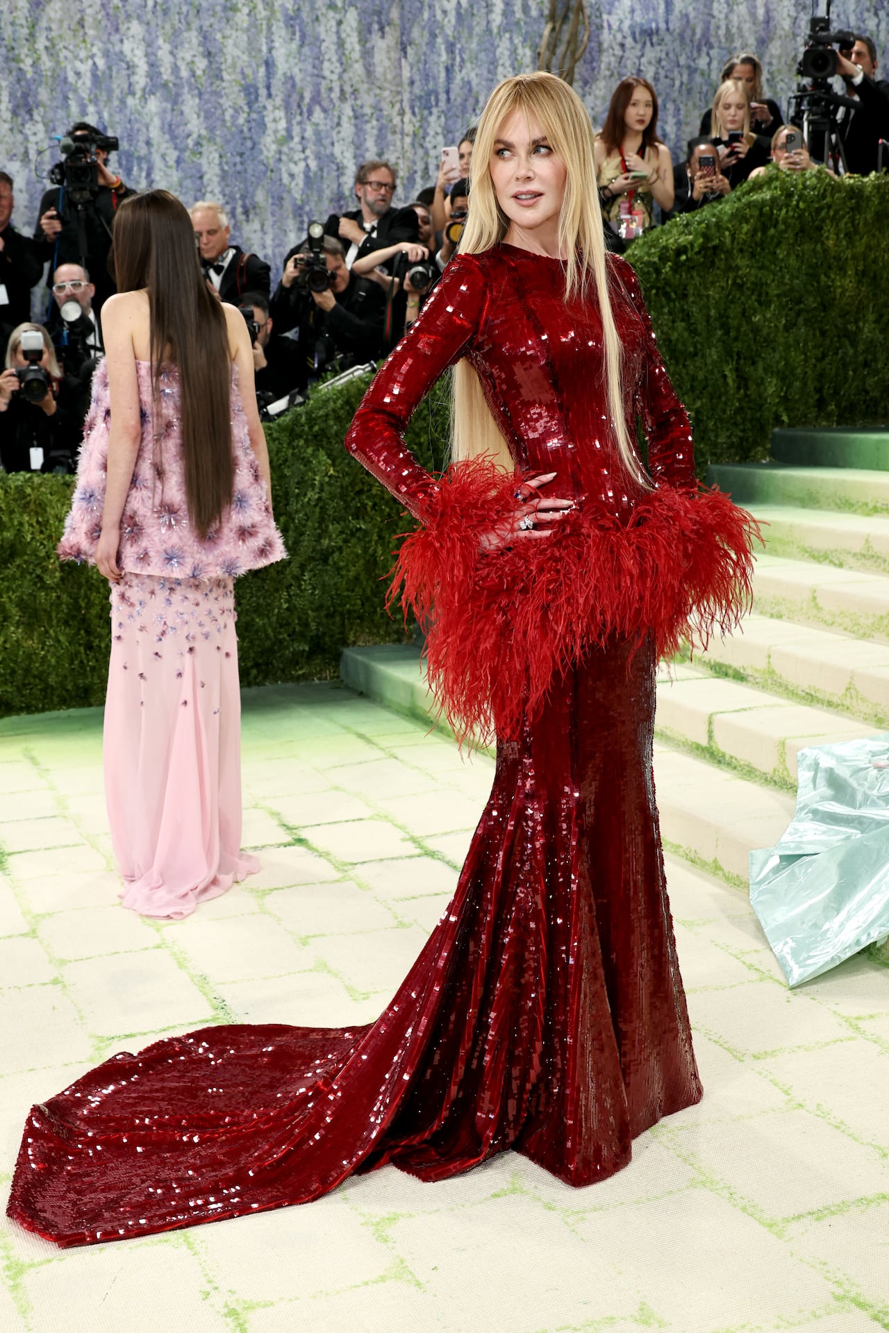 A woman wearing a long red sequinned gown with a train. A layer of long, thin feathers sits at the waist.