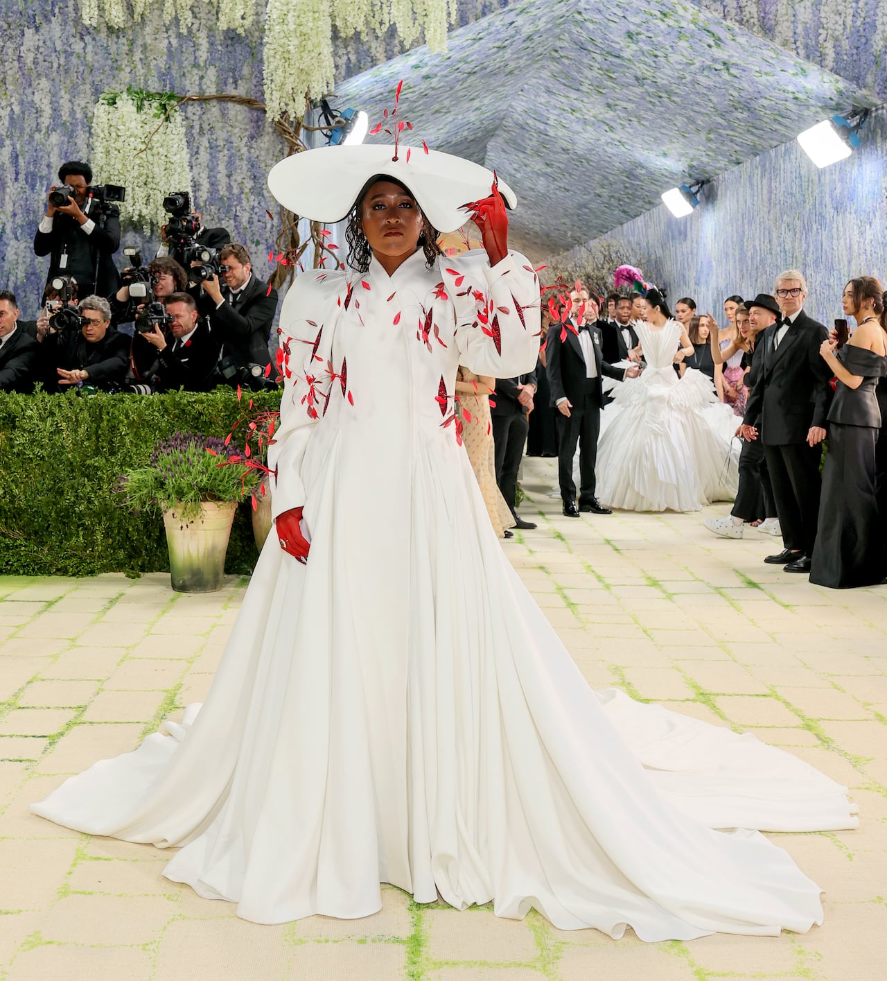 A woman poses for a photo wearing a long sleeve voluminous white gown with some red feathers on it and a large wide-brimmed hat.