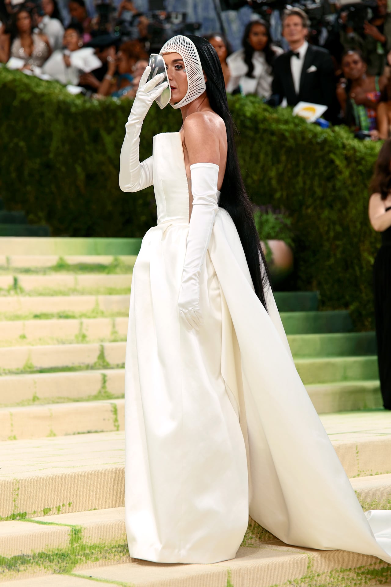 A woman poses for a photo wearing a strapless cream gown and lifts a silver mask to reveal her face.