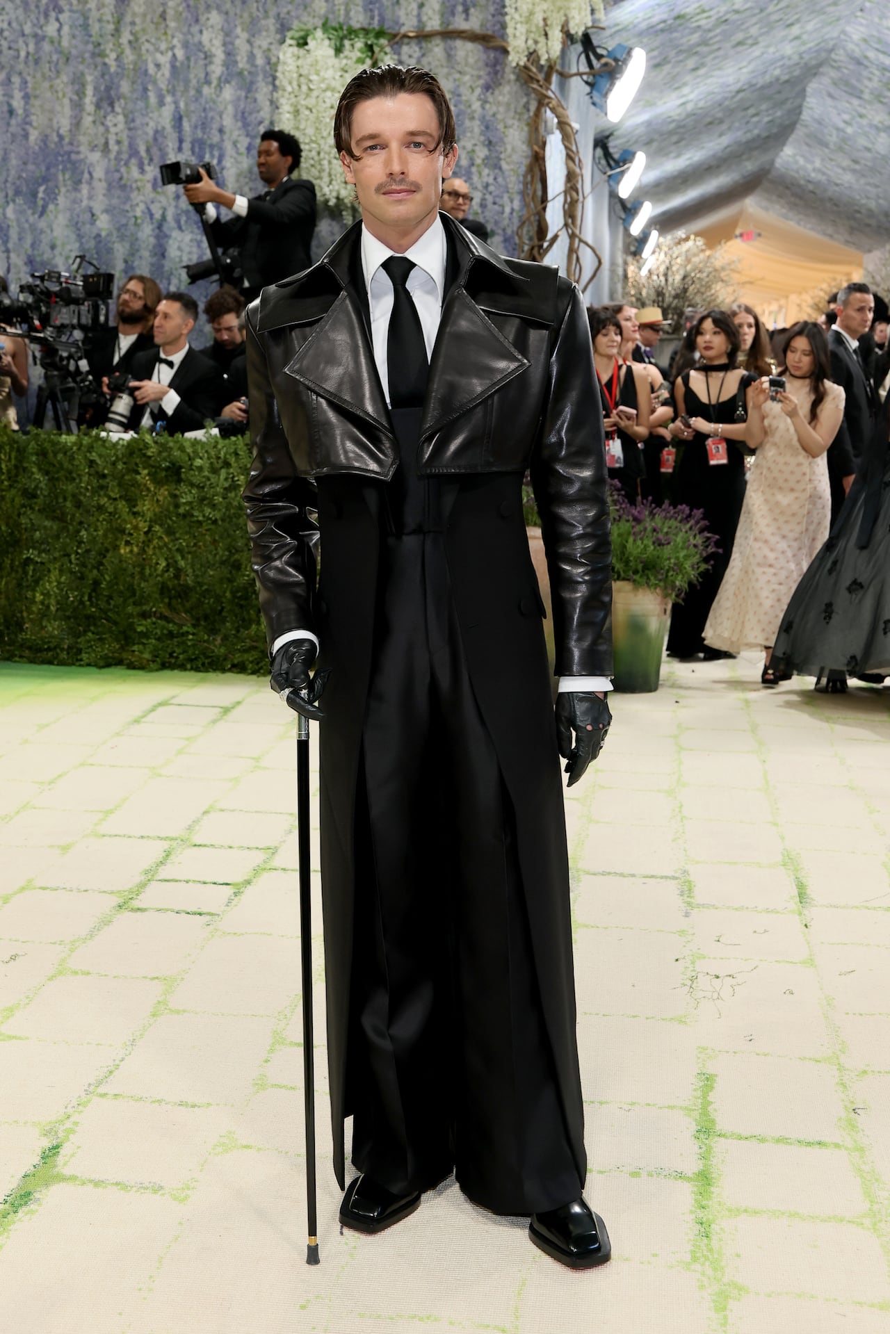 A man poses for a photo wearing a black suit with a cropped leather jacket over top and he's carrying a cane.