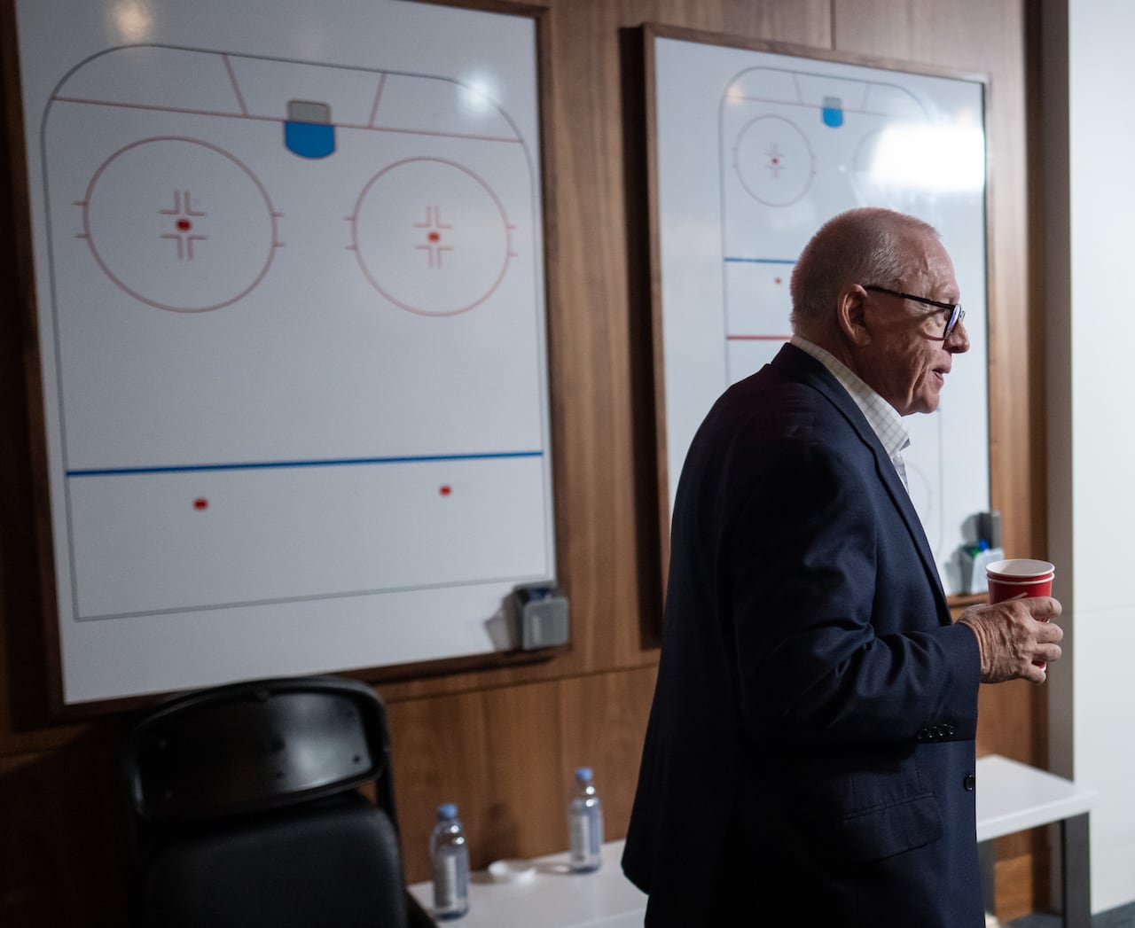 A white man carrying coffee cups walks past two whiteboards with hockey diagrams on them.