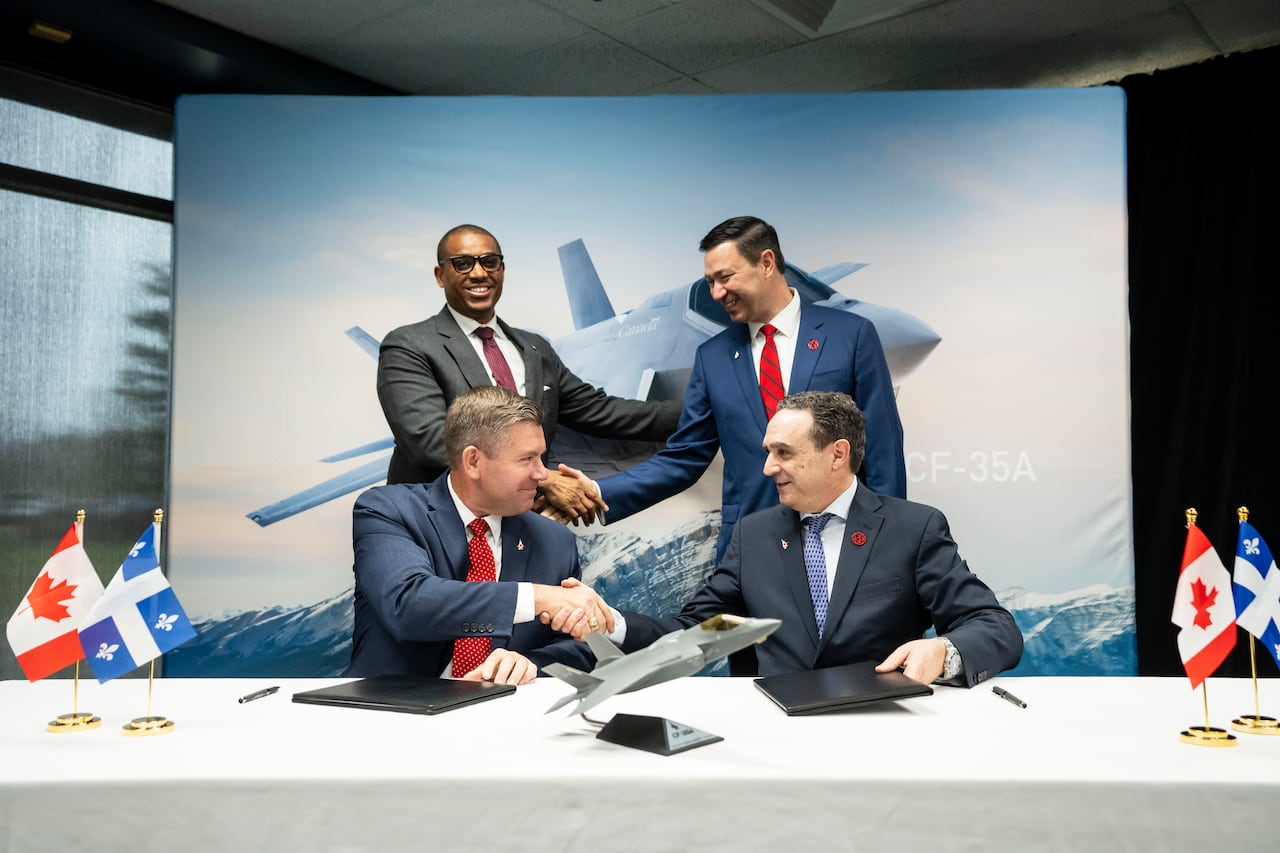 Four men shake hands in front of a picture of a plane.