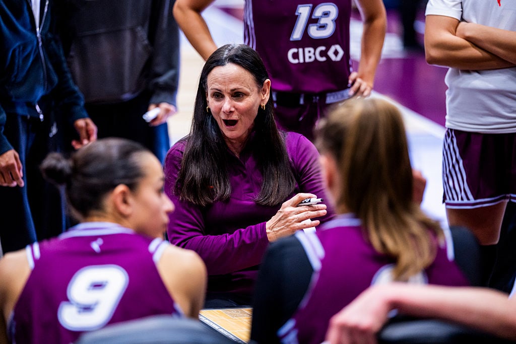 A women's basketball coach is shown crouching to speak with players.
