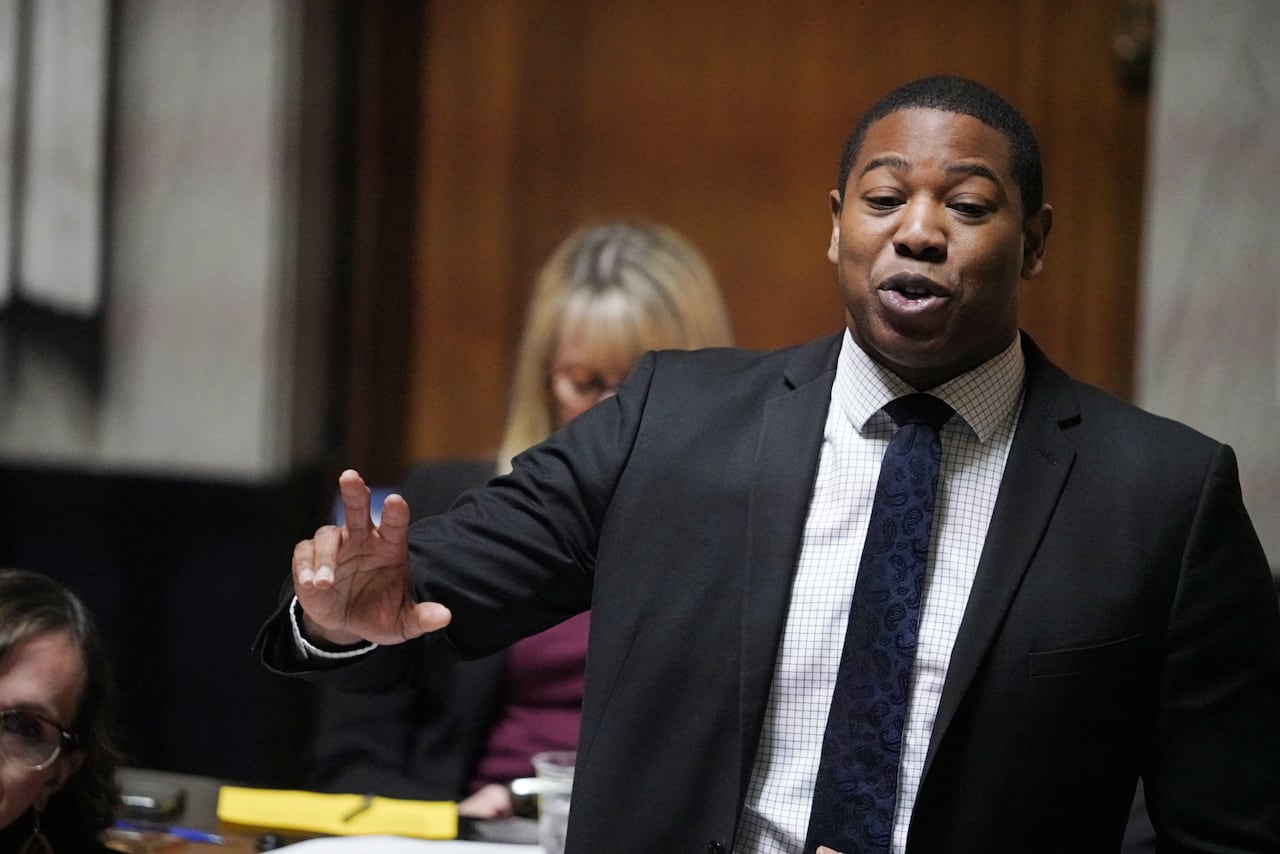 A man in a black suit and white patterned shirt gestures with his right arm while speaking during a government legislative session.