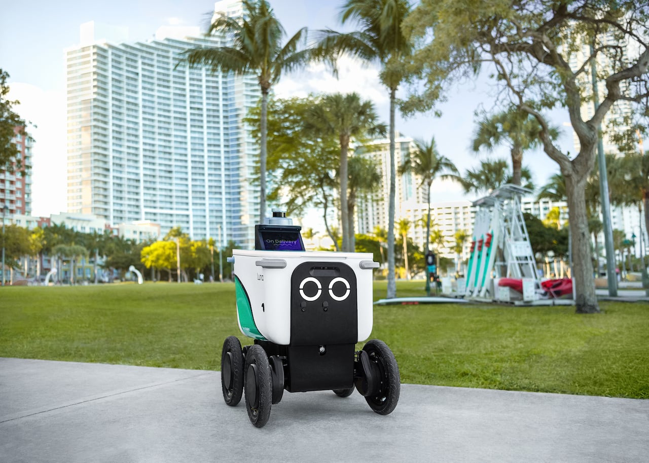 A small cube-shaped robot is seen on a sidewalk on a sunny day.