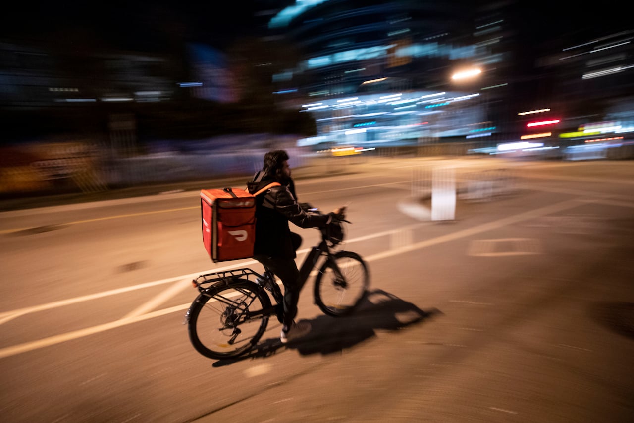 A cyclist with an orange DoorDash delivery bag bikes at night down a Vancouver street.