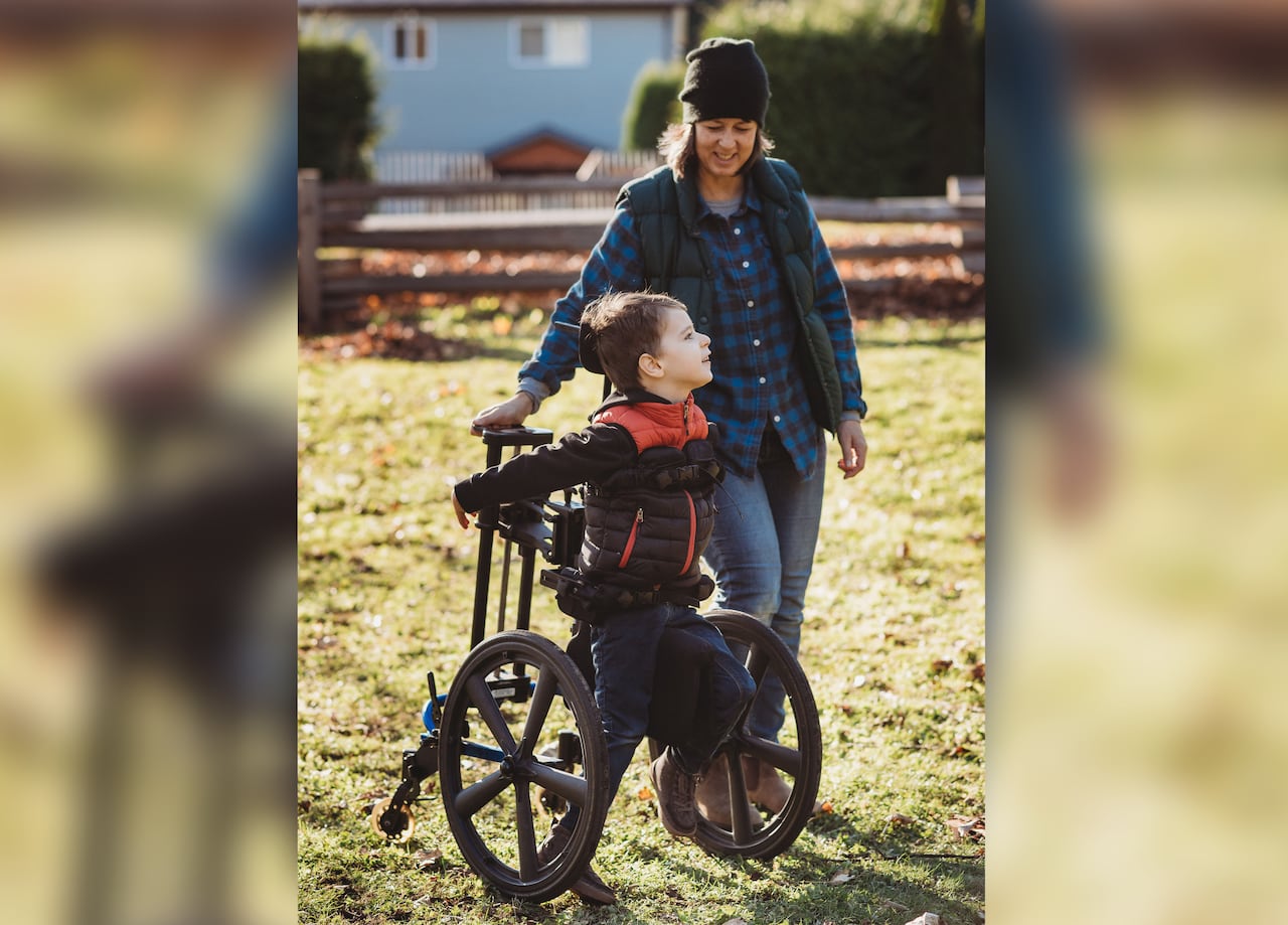 A young boy in a wheelchair is pictured on a grassy field with a woman standing next to him, both of them are smiling.